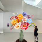 A person standing next to a large, colorful, freestanding artificial flower bouquet with metal bases.